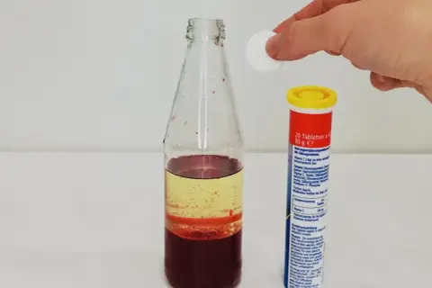 Eine Hand lässt eine Tablette in eine Flasche mit bunten Flüssigkeiten fallen, um eine Lavalampe selber zu machen.