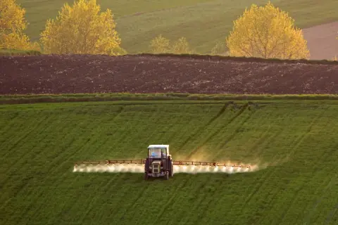 Traktor wykonuje oprysk na zielonym polu. W tle widać zaoraną ziemię i żółte drzewa.
