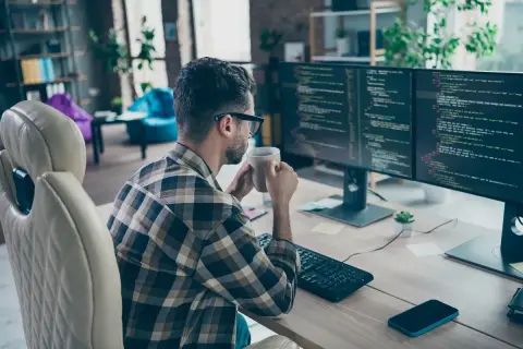 Programista w okularach pije kawę, pracując zdalnie przy dwóch monitorach z kodem.