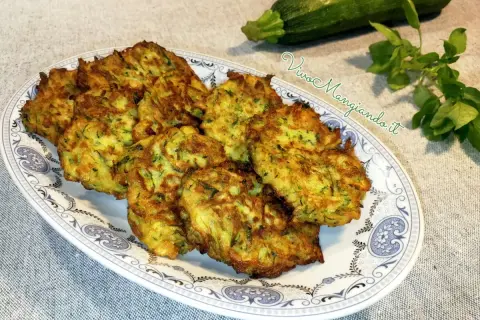 Frittelle di zucchine dorate e croccanti, servite su un piatto decorato. Accanto, una zucchina fresca e un rametto di erbe aromatiche.