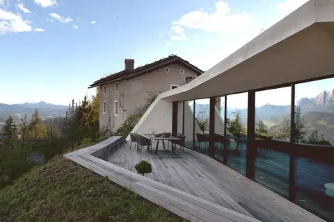 Modernes Haus in Hanglage mit Panoramafenstern und Terrasse, daneben ein altes Bauernhaus, umgeben von Berglandschaft.