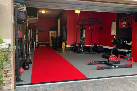 A fully equipped garage gym with a red carpet runner, weight racks, a punching bag, and a "Stronger 40" banner, ready for some serious garage boxing.