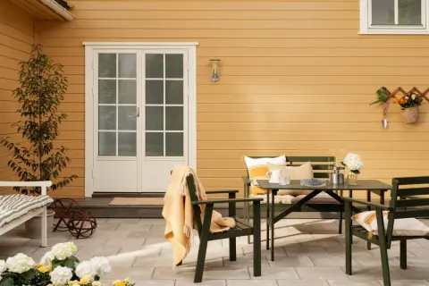Ett gult hus med svart tak. En uteplats med gröna möbler, blommor och en orange pläd.
