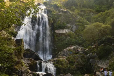 La majestuosa cascada de Belelle cae en cascada por rocas cubiertas de musgo, con gente disfrutando del paisaje y el agua.