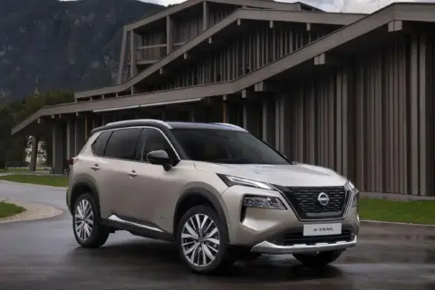 Elegancki, beżowy Nissan X-Trail 2015 zaparkowany przed nowoczesnym budynkiem w górach.