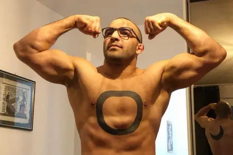 Muskularny mężczyzna o arabskich rysach, w okularach, prezentuje swoje bicepsy. Na klatce piersiowej ma tatuaż w kształcie pierścienia.