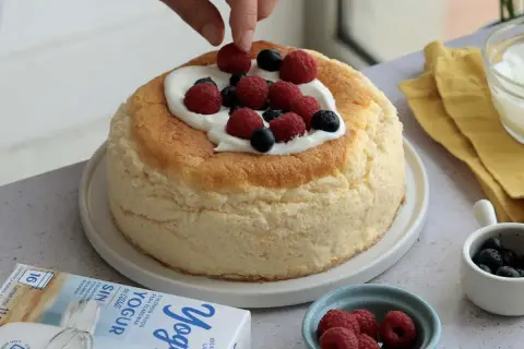 Tarta de queso esponjosa decorada con frutos rojos y nata. Se ve una caja de yogur griego al lado.