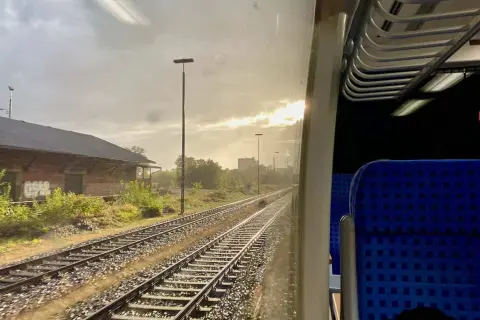 Blick aus dem Zugfenster auf Gleise und ein altes Backsteingebäude. Die Sonne bricht durch die Wolken. Man fragt sich, wie viel kostet eine Zugfahrt.