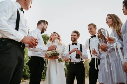 Świadek na weselu otwiera szampana, podczas gdy panna młoda i pan młody cieszą się chwilą z przyjaciółmi.