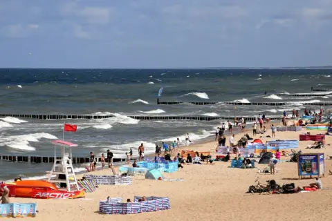 Plaża nad Bałtykiem pełna ludzi, fal i parawanów. Widać ratownika, sprzęt wodny i tablicę informacyjną. Ceny nad morzem pewnie wysokie, ale widok wart zachodu.