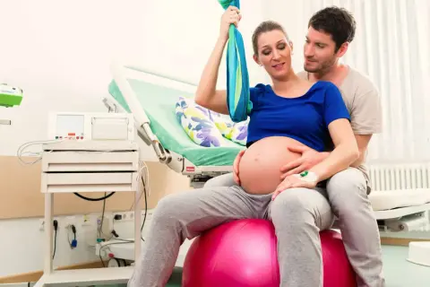 Pareja en sala de partos. Ella sentada en balón, buscando la mejor postura para parir, él la abraza.
