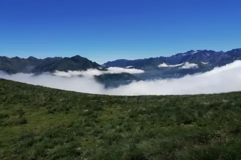 Vue panoramique depuis le col de Riou, avec des montagnes verdoyantes émergeant d'une mer de nuages sous un ciel bleu éclatant.