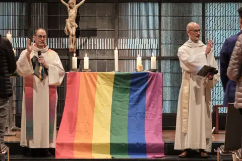 Zwei Geistliche stehen vor einem Altar mit Regenbogenflagge. Ein Kreuz hängt im Hintergrund. Die Kirche feiert die gleichgeschlechtliche Ehe.