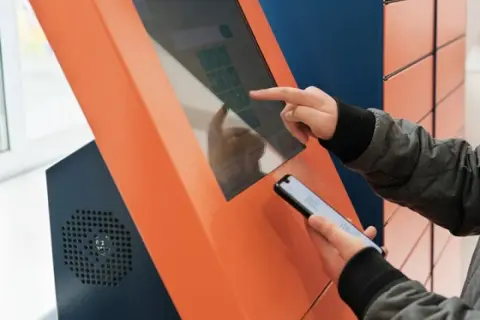 Osoba korzysta z ekranu dotykowego automatu, by dowiedzieć się, jak wygenerować etykietę InPost. Trzyma też telefon.