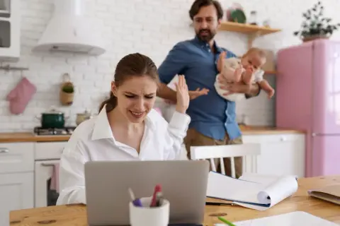 Zestresowana kobieta przy laptopie, mąż z dzieckiem w tle. Czy przerwa na karmienie piersią do jakiego wieku dziecka jest możliwa w takiej sytuacji?