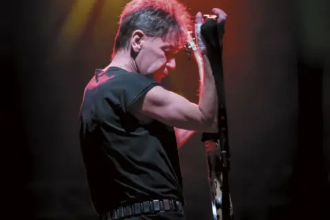 Muzyk w czerni, z czerwoną poświatą na włosach, przygotowuje się do wykonania swoich ksu utworów na gitarze.