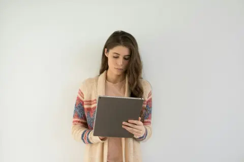 Młoda kobieta w kardiganie z wzorami przegląda tablet, szukając ofert pracy dla psychologa online.