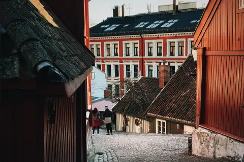 Två personer promenerar på en kullerstensgata i Oslo centrum, omgivna av charmiga trähus och en ståtlig röd byggnad.