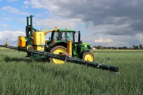 Zielony traktor z opryskiwaczem polowym, gotowy do pracy z adiuwantem, na tle pola i chmur.