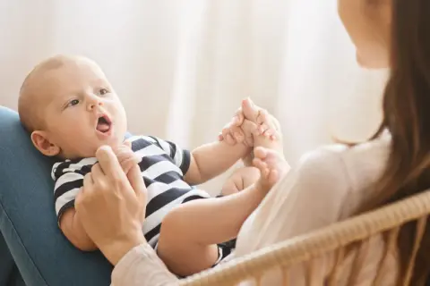 Ein Baby mit offenem Mund liegt auf dem Schoß seiner Mutter. Es hält ihre Hände. Bald sagt das Baby "Mama".