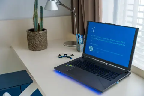 Laptop z niebieskim ekranem śmierci, wymagający twardy reset laptopa. Na biurku okulary i kaktus.