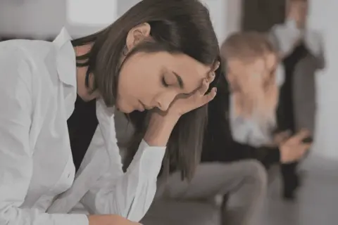 En ung kvinna med handen mot pannan, kanske i väntan på en **social fobi diagnos**. I bakgrunden syns andra personer.