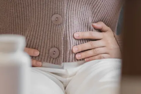 Kobieta trzyma się za brzuch, szukając ulgi na wzdęcia. Obok stoi biała butelka z lekami.