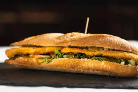 Un bocadillo caliente con rebozado crujiente, lechuga fresca y pepinillos, servido en pan rústico.