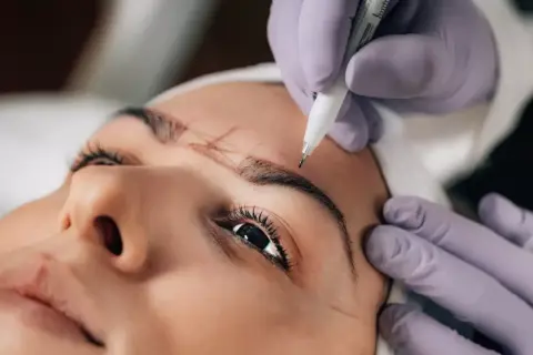 Manos enguantadas dibujan el contorno de las cejas para un procedimiento de powder brows.
