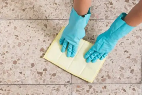 Manos con guantes azules limpiando suelos de terrazo con un paño amarillo.