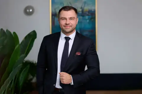 Mężczyzna w garniturze, stojący obok rośliny, z obrazem portowym w tle.