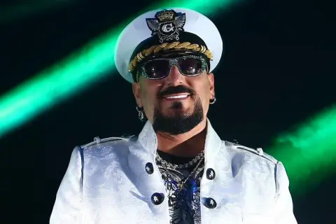 Gigi D'Agostino w białej marynarce i czapce, uśmiechnięty. Mimo choroby, jego muzyka wciąż porusza serca fanów.