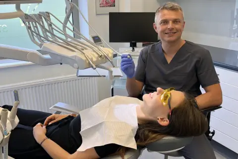Dentysta w rękawiczce trzyma wiertło, pacjentka w okularach ochronnych leży na fotelu.