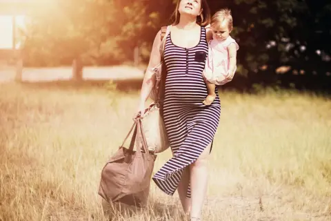 Schwangere Frau mit Baby im Arm und Taschen, die sich durch hohes Gras kämpft. Schwer heben schwangerschaft.