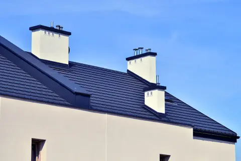 Nowoczesny dach bezokapowy z ciemnogranatową dachówką i białymi kominami, pod błękitnym niebem.