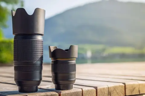 Two camera lenses, a tall telephoto and a shorter normal lens, sit on a wooden deck with a blurred mountain and lake background.
