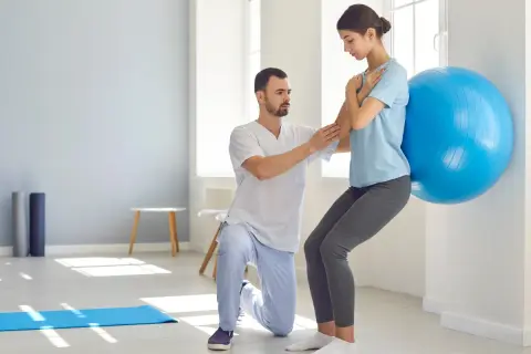 Fizjoterapeuta pomaga kobiecie w ćwiczeniu aktywującym mięśnie głębokie przy użyciu piłki gimnastycznej.