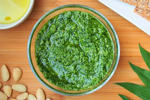 Aromatic pesto z czosnku niedźwiedziego w szklanej miseczce, otoczone migdałami i świeżymi liśćmi.