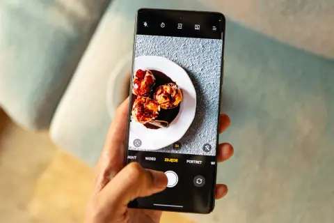 Dłoń trzyma smartfon z otwartą aplikacją do robienia zdjęć. Na ekranie widoczny jest talerz z trzema ptysiami polanymi czekoladą.