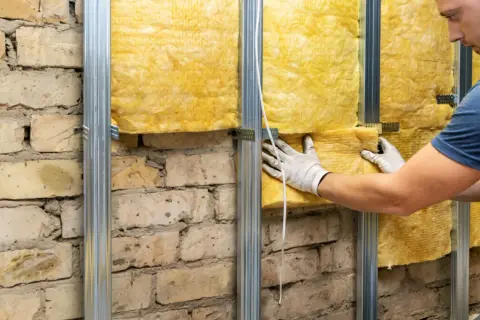 Pracownik w białych rękawicach montuje wełnę mineralną na stelażu, pokazując jak ocieplić fundamenty starego domu.