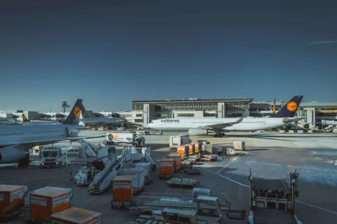 Samolot Lufthansa na płycie lotniska, gotowy do przyjęcia bagażu podręcznego. W tle budynek terminala i inne samoloty.