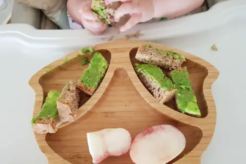 Babyhände greifen nach Avocado-Brotstücken auf einem Holzbrett. Ein 10 Monate altes Baby erkundet sein Essen.