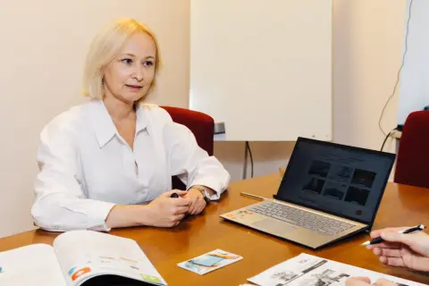 Kobieta w białej koszuli uczy się słówek po rosyjsku, korzystając z laptopa i materiałów drukowanych.