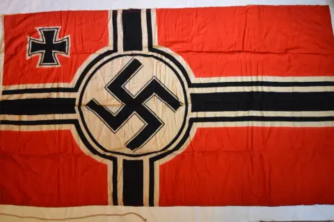 Drapeau rouge avec une croix noire et blanche dans le coin supérieur gauche et une croix gammée noire sur un cercle blanc au centre. L'origine de la croix gammée est complexe.
