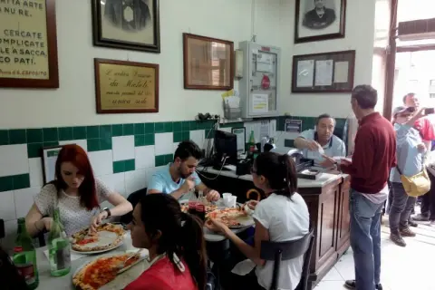 Wnętrze najstarszej pizzerii w Neapolu. Ludzie jedzą pizzę, a za ladą obsługuje kelner.