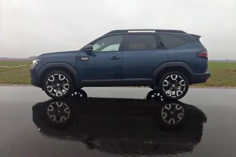 Dacia Bigster LPG w ciemnoniebieskim kolorze, zaparkowana na mokrej drodze, z odbiciem w kałuży.