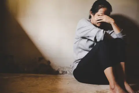 Kobieta siedzi skulona w kącie, zakrywając twarz dłońmi. Ciemność i światło podkreślają jej rozpacz, być może spowodowaną uzależnieniem od kokainy.