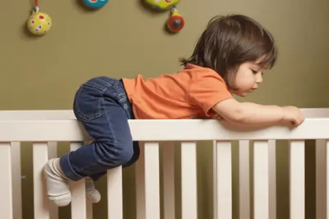A toddler in jeans and an orange shirt is climbing over the side of a crib, with their legs dangling, as if preparing for dropping crib mattress to floor.