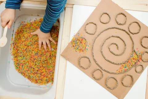 Main colorée dans un bac, une main d'enfant explore. À côté, un carton en spirale avec des ronds découpés, parsemé de riz coloré. Une activité sensorielle adulte pour stimuler la créativité.