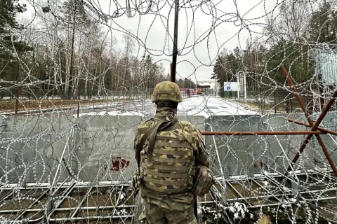 Żołnierz w mundurze obserwuje teren przez zasieki z drutu kolczastego. Widoczna jest granica polsko-białoruska, z budynkami i znakami po drugiej stronie.